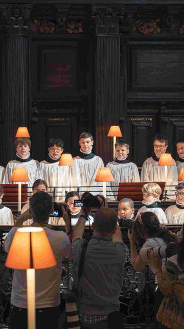 St Paul's Cathedral choristers stand in the Cathedral quire, which is lit with lamps. They are stood in two rows, and are being photographed by a group of press photographers.