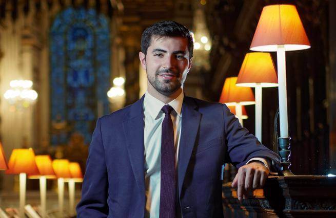 James is a white man with short dark hair wearing a blue suit. He stands in the quire at St Paul's Cathedral, leaning against a stall, with lit lamps alongside.