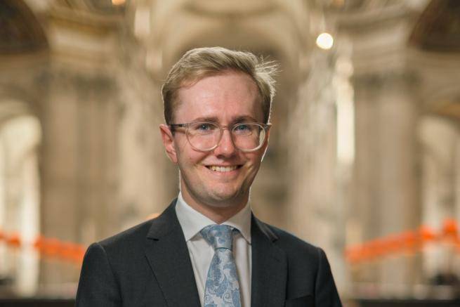 Hamish Wagstaff smiling, wearing glasses, dark suit, and light blue tie with floral pattern