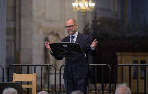 Allen stands at a lectern on a stage in St Paul's Cathedral, arms out in front of him as he speaks to an audience