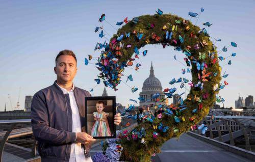 A man stands on Millennium Bridge in London, dressed in a white shirt and blue jacket, with short blond hair. He is holding a photo of his late daughter, who passed away from cancer. He is standing beside an art work for Great Ormond Street Hospital Charity, made of butterflies, framing the dome of St Paul's Cathedral.