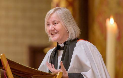 Ellen wears a clerical collar and robes while standing at a lectern, her hand outstretched as she talks
