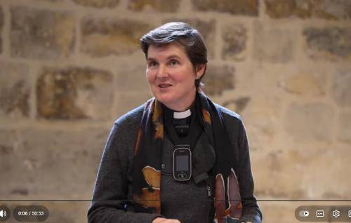 A still from the film in which Morwenna stands in the Wren Suite speaking to an audience